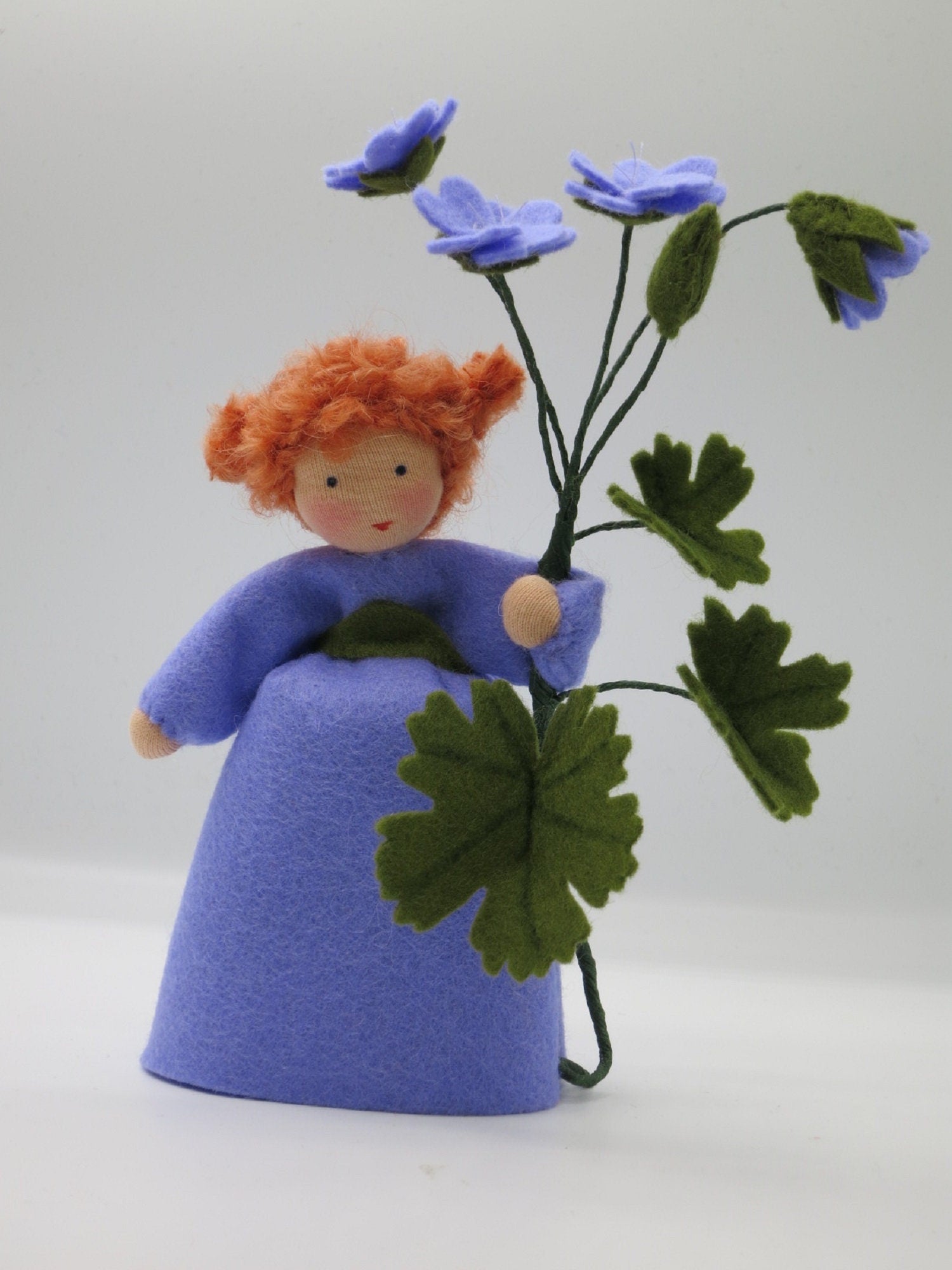 Storchschnabel-Blumenkind aus Wollfilz für den Waldorf Jahreszeitentisch mit drei violetten Blüten und gelappten Blättern.